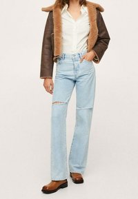 Femme portant une veste en peau retournée marron, une chemise blanche boutonnée, un jean bleu clair déchiré et des bottines marron, debout avec une main dans la poche.