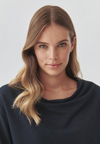 Portrait einer Person mit welligem, schulterlangem blonden Haar, die ein marineblaues, drapiertes Oberteil mit Rundhalsausschnitt trägt. Glatte Haut, neutrale Ausdruck.