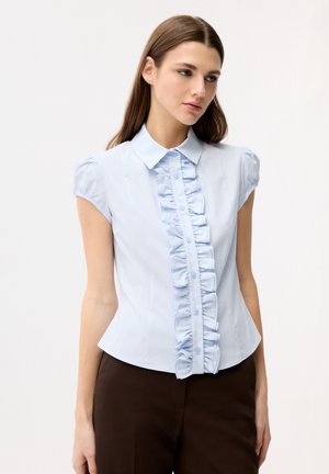 Ung kvinde iført en lyseblå kortærmet bluse med flæsedetaljer foran og mørke bukser, stående mod en ensfarvet baggrund.