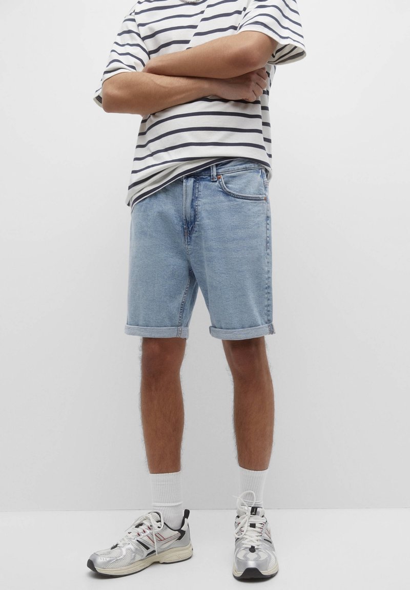 Shorts en denim bleu clair avec des ourlets retroussés, associés à un T-shirt rayé blanc et marine. Portés avec des baskets métalliques et des chaussettes blanches.