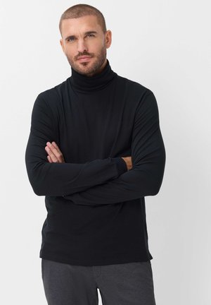 Uomo con capelli corti e barba che indossa un dolcevita nero e pantaloni scuri, in piedi con le braccia incrociate su sfondo bianco.