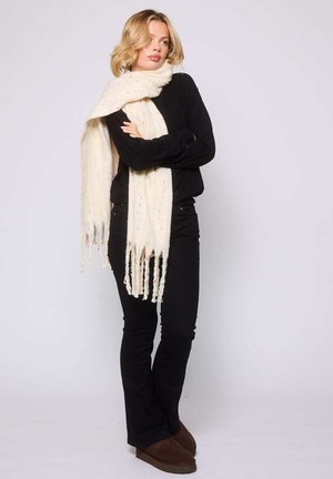Femme blonde portant une tenue noire, une écharpe crème surdimensionnée avec des franges et des chaussures marron, se tenant les bras croisés sur un fond uni.
