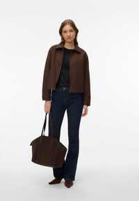 Femme portant une veste marron, un haut noir, un jean bleu foncé et des chaussures marron, tenant un grand sac fourre-tout marron sur un fond blanc.