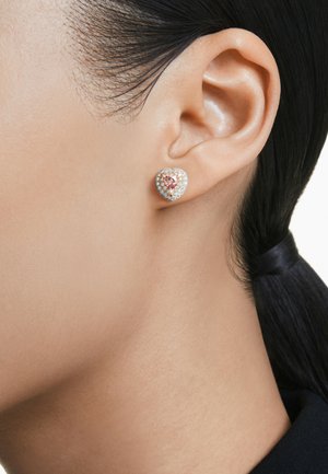 Hartvormige oorbel met een roze edelsteen in het midden, omringd door een cirkel van kleine witte steentjes; metalen afwerking, gladde textuur.