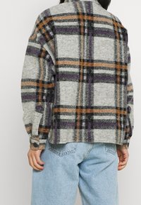 Grijs geruit flanellen overhemd met paarse, oranje en zwarte strepen, met een kraag en verstelbare manchetten, gedragen over lichtblauwe spijkerbroek.