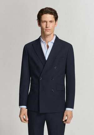 Hombre con cabello castaño corto que lleva una chaqueta de traje cruzada de color azul marino oscuro y una camisa de vestir azul claro, de pie frente a un fondo liso.