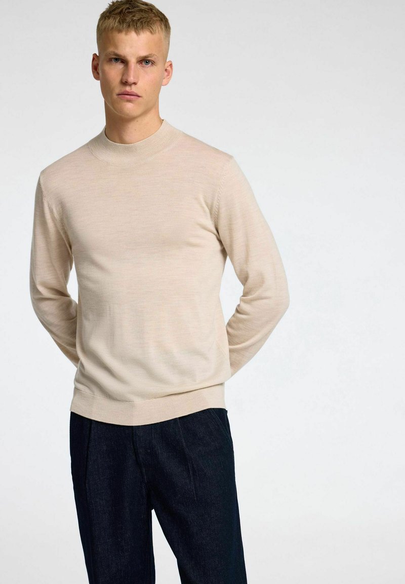 Pullover in maglia beige con scollatura tonda, maniche lunghe e orlo a costine. Il tessuto ha una consistenza liscia. Indossato con pantaloni scuri.
