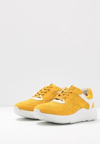 Pair of yellow suede sneakers with white accents and thick white soles placed on a white surface against a light gray background.