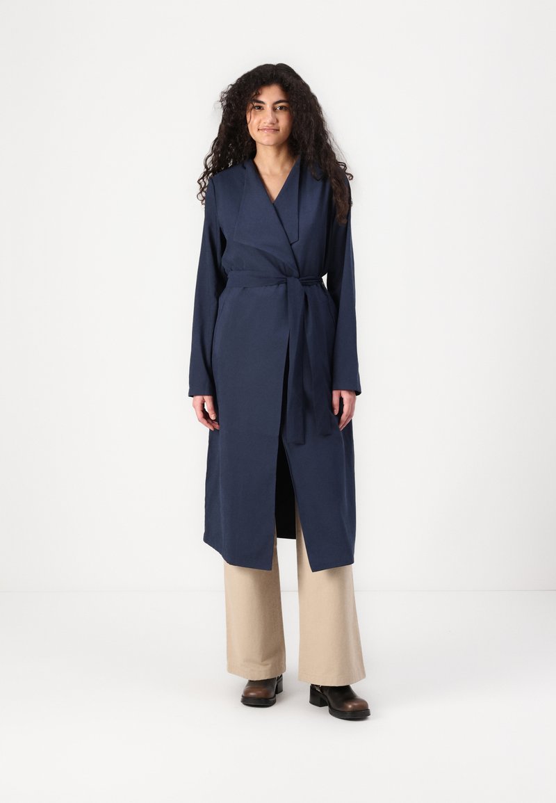 Navy blue long wrap dress with a tie belt, long sleeves, and a V-neck. Paired with beige wide-leg pants and dark brown block-heeled boots.