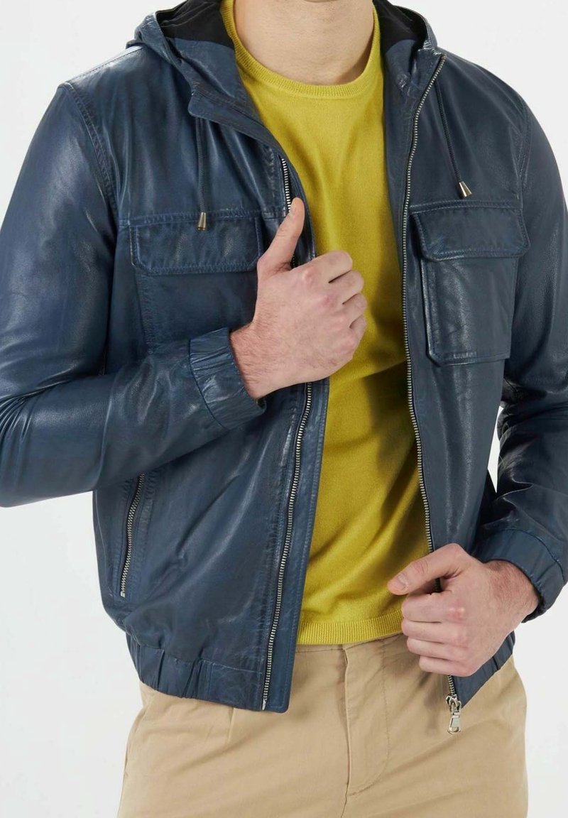 Homme portant une veste en cuir bleu marine sur un pull jaune moutarde, ajustant la fermeture éclair de la veste avec les mains.