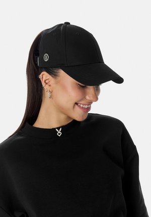 Kvinde med mørkt hår samlet i en hestehale bærer flere øreringe, en sort kasket med et lille logo og en sort skjorte med et kaninhjerteemblem.