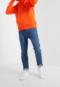 Orange huvtröja med framficka, tillsammans med uppvikta blåjeans och vita sneakers. Rena linjer och enkel design.