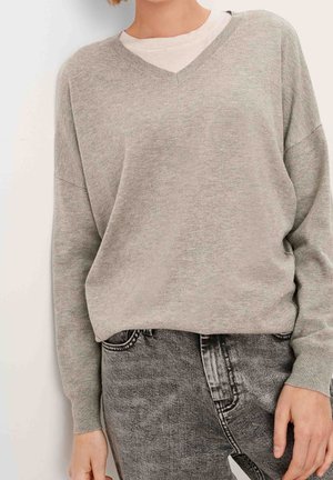 Personne portant un pull à col en V gris clair sur un t-shirt blanc, associé à un jean noir délavé taille haute.
