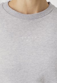 Lysegrå treningsoverdel med ribbestrikket rund hals, med brodert "HYROX"-logo. Materialet ser mykt og teksturert ut.