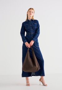 Denimska haljina s prednjim kopčanjem na dugmad, dugim rukavima i dva džepa na grudima. Model drži smeđu torbu od mekog materijala, noseći sandale s trakama.