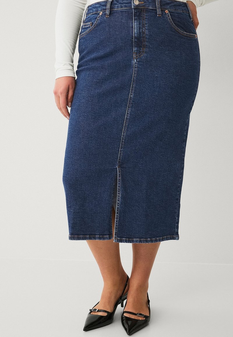 Jupe midi en denim bleu foncé avec une fente à l'avant, design à cinq poches, passants de ceinture et couture contrastée. Portée avec des talons noirs à bout pointu.