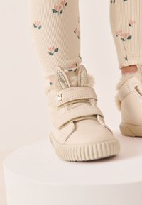 Beige velcro sneakers with rabbit ear accents, furry trim, and a textured rubber sole. Worn with cream ribbed socks featuring floral patterns.