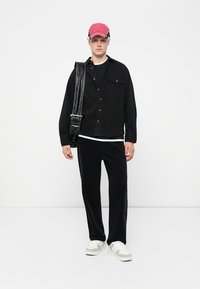 Black button-up shirt over a black t-shirt, black corduroy pants, white sneakers with gray accents, and a red cap with a graphic design.