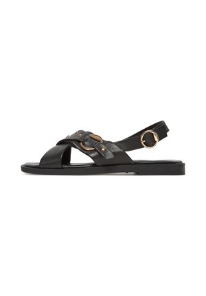 Sort flad sandal med krydsede remme, guldmetaldetaljer og en justerbar ankelrem med en rund guldbøjle.