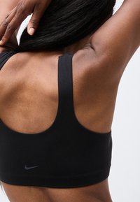 Prim-plan al unei persoane care se întinde cu brațele ridicate, purtând un sutien sport negru Nike, care arată spatele tonifiat și pielea netedă.