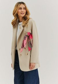 Beige dvourohý blazer s texturovaným povrchem, kombinovaný s bílým topem a tmavými džíny. U kapsy je uvázaný barevný šál s potiskem.