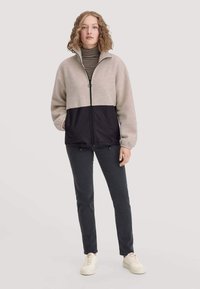 Fleecejacke mit einem hellbeigen oberen und einem dunkelblauen unteren Bereich, ausgestattet mit einem Reißverschluss und hohem Kragen. Kombiniert mit dunkler Hose und Turnschuhen.