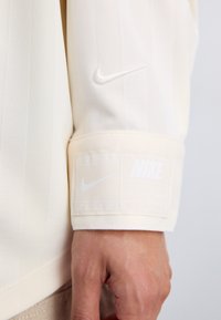 Bijela, teksturirana sportska odjeća s Nike logom na rukavu i etiketom s imenom brenda i uputama za njegu.