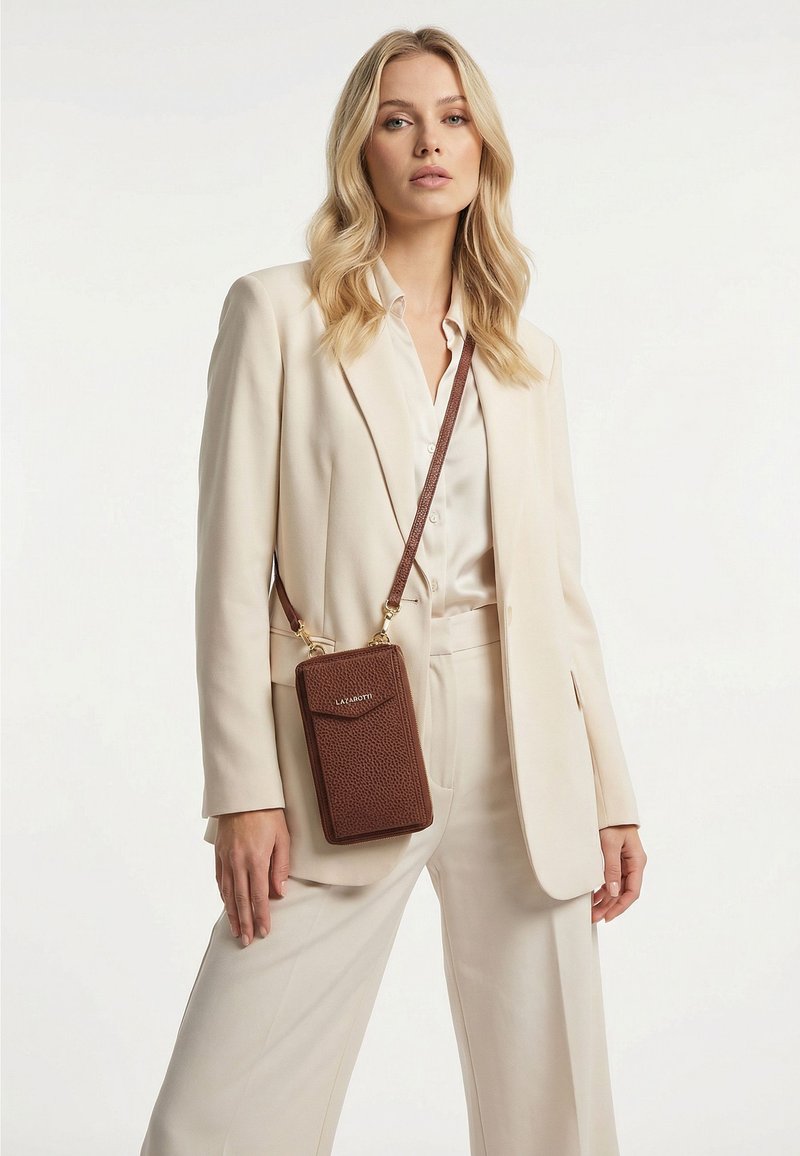 Mujer rubia con traje crema y blusa blanca lleva un bolso bandolera marrón con textura, herrajes dorados y una correa cruzada sobre el pecho.