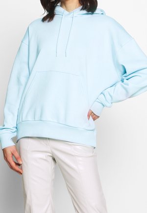 Osoba nosząca jasnoniebieską, oversize'ową bluzę z kapturem i kremowe skórzane spodnie, pozująca z jedną ręką na biodrze, a drugą swobodnie opuszczoną.