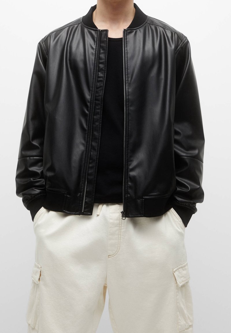 Personne portant une veste bomber en cuir noire sur un t-shirt noir avec un pantalon cargo blanc, mains dans les poches, sur fond uni.