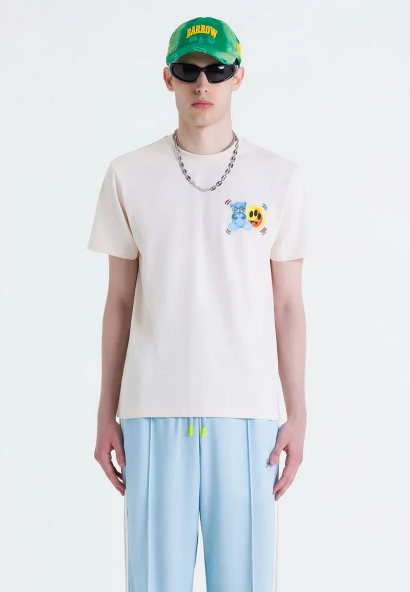 Camiseta de algodón color crema con un gráfico de un oso azul y una carita sonriente, combinada con pantalones azul claro con acentos neón y franjas laterales. Gafas de sol negras y gorra verde.