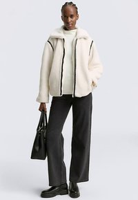 Džemper od flisa u nijansi prljavo bijele boje s prednjim patentnim zatvaračem, crnim obrubom i visokim ovratnikom. Kombiniran s krem džemperom, crnim širokim hlačama i čizmama.