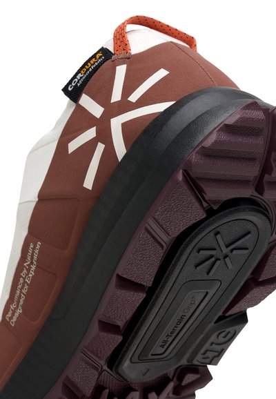 Gros plan sur une chaussure de randonnée marron et blanche montrant la semelle crantée avec "Adhérence Tout-Terrain" et une languette avec l'étiquette Corouf.