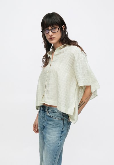 Ανοιχτό πράσινο πουκάμισο με κοντά μανίκια και κουμπιά, με υφασμένη υφή, φορεμένο πάνω από ψηλόμεσα μπλε τζιν. Διαθέτει άνετη εφαρμογή και γιακά.