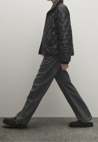 Osoba idąca, ubrana w czarną pikowaną kurtkę, luźne ciemnoszare d�żinsy oraz czarne buty wsuwane, na szarej podłodze na tle jednolitej ściany.