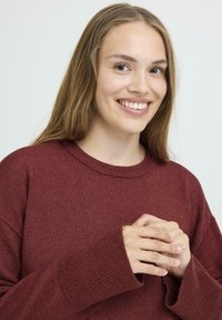 Pull en maille bordeaux avec un col rond et des épaules tombantes. Présente des poignets côtelés et un tissu texturé. Le modèle a les mains jointes.