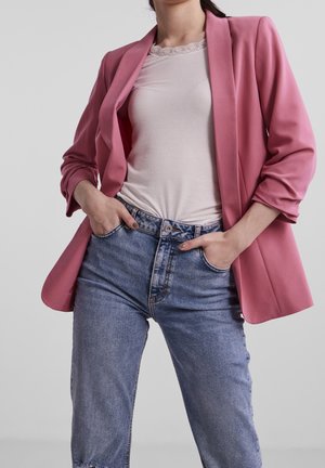 Femme portant un blazer rose, un haut blanc avec bordure en dentelle, et un jean taille haute bleu, les mains dans les poches, debout devant un fond uni.