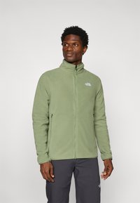 Veste en polaire verte avec un col haut, fermeture éclair à l'avant et deux poches zippées. Présente un petit logo blanc sur la poitrine gauche.