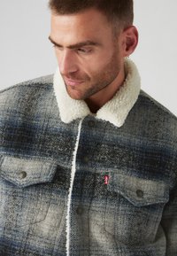 Chaqueta de mezcla de lana con un patrón de cuadros en tonos de azul y gris, con un cuello de sherpa color crema y bolsillos frontales con botón.