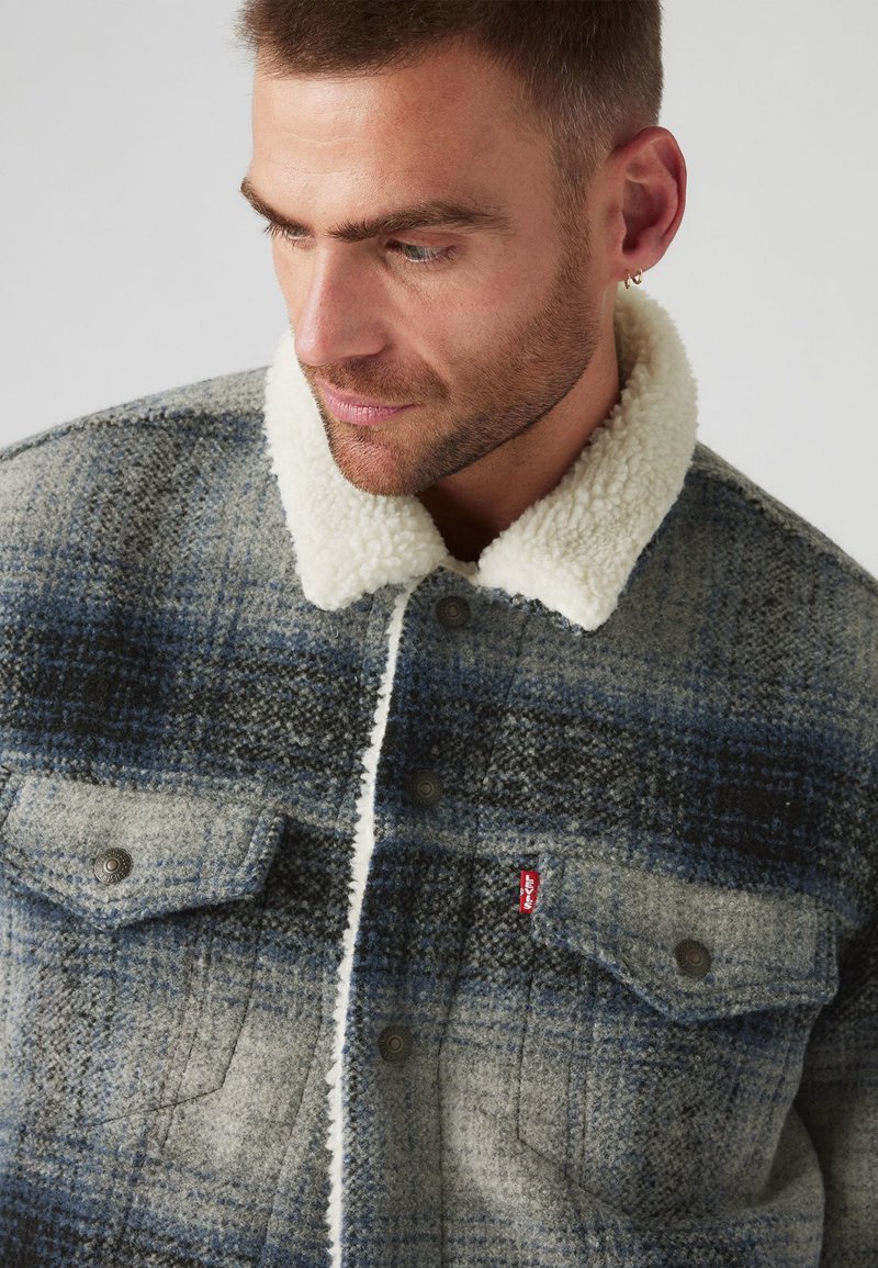 Veste en mélange de laine avec un motif à carreaux dans des tons de bleu et de gris, dotée d'un col en sherpa de couleur crème et de poches à bouton à l'avant.