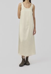 Robe maxi sans manches de couleur crème en tissu lisse, avec un col rond. Associée à des bottines marron.