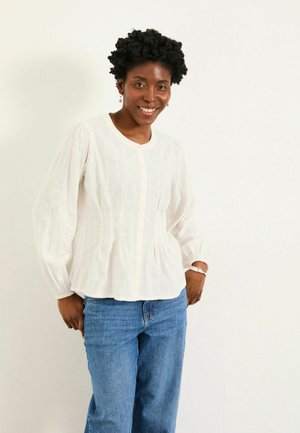 Lächelnde Frau mit kurzen lockigen Haaren, die eine weiße Langarmbluse und blaue Jeans trägt und vor einer schlichten weißen Wand steht.