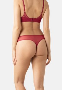 Ensemble de lingerie rouge comprenant un bralette transparent avec un dos à motifs et un string assorti, tous deux avec des textures lisses et des bretelles ajustables.
