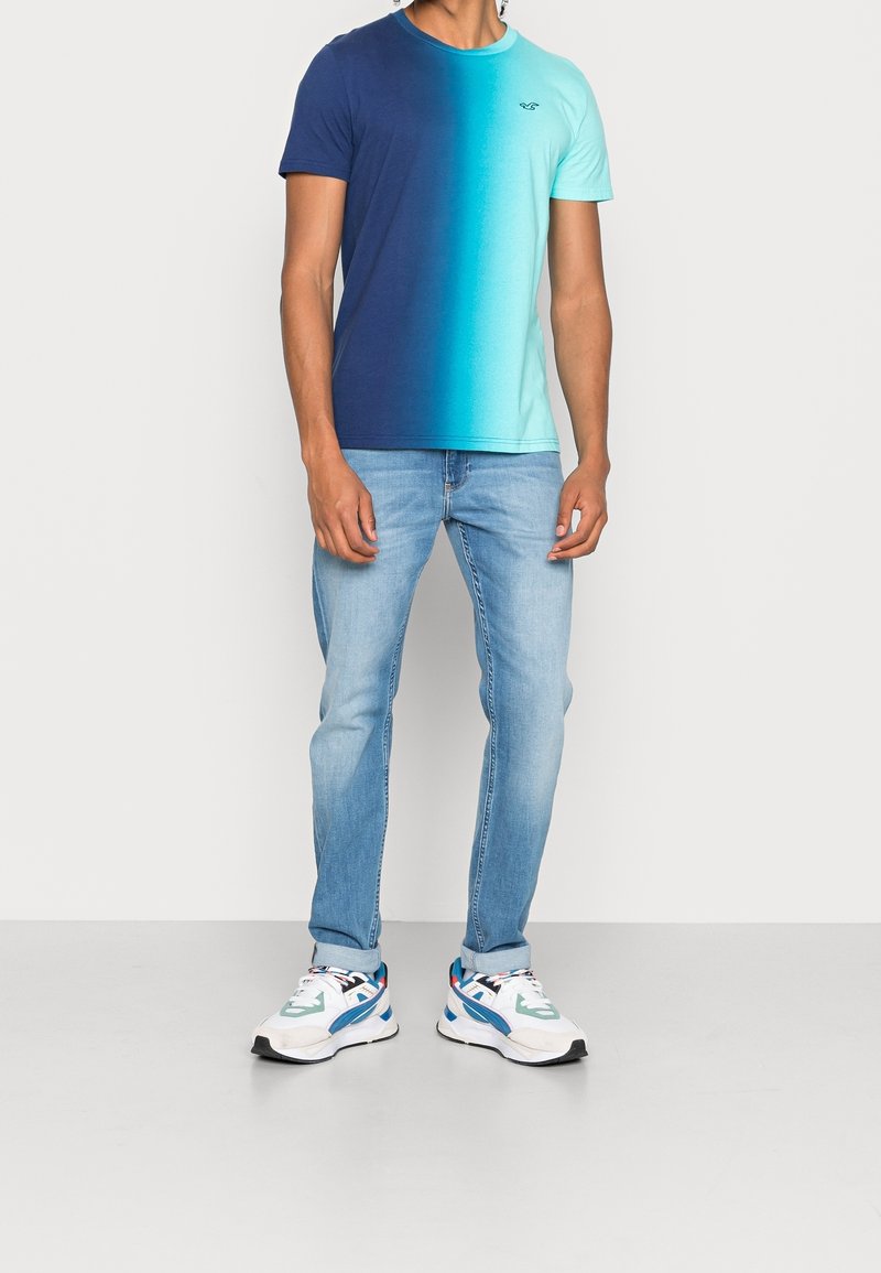 Man in een gradient marineblauw tot turquoise T-shirt, lichtblauwe jeans met opgerolde broekspijpen en witte sneakers met blauwe en turquoise accenten.
