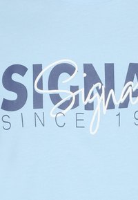 Šviesiai mėlynos medvilnės marškinėliai su ryškiai tamsiai mėlyna užrašu "SIGN" ir dekoratyviniu "a", papuoštu baltais akcentais, pabrėžiančiu prekės ženklą ir metus.