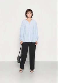 Blusa azul clara de botão com mangas compridas e cintura franzida, combinada com calças largas pretas e sandálias pretas. Pequena mala preta.