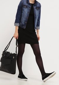 Svart ribbat klänning, jeansjacka med uppvikta ärmar, svarta sneakers och strukturerad svart handväska. Modell går, visar upp outfitdetaljer.