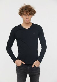 Chemise noire à manches longues en V avec texture côtelée, comportant un petit logo sur la poitrine. Associée à un jean foncé.