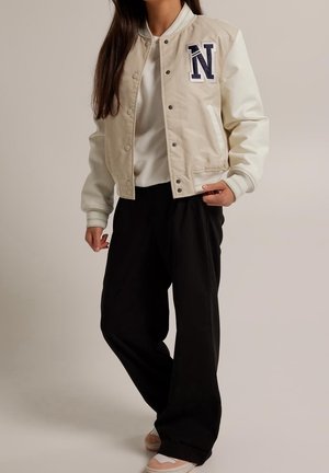 Donna che indossa una giacca varsity beige e bianca con toppa "N" blu navy, camicia bianca, pantaloni neri a palazzo e sneakers rosa e bianche.