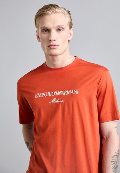 T-shirt en coton rouge avec un col rond. Présente un logo blanc brodé "Emporio Armani Milano" sur la poitrine. Manches courtes, texture lisse.